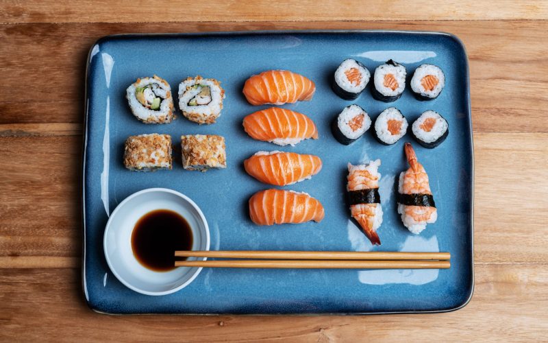 sushi-plate-with-maki-uramaki-nigiri-california-roll-blue-plate-soy-chopsticks-asian-gastronomic-food-composition-wooden-background